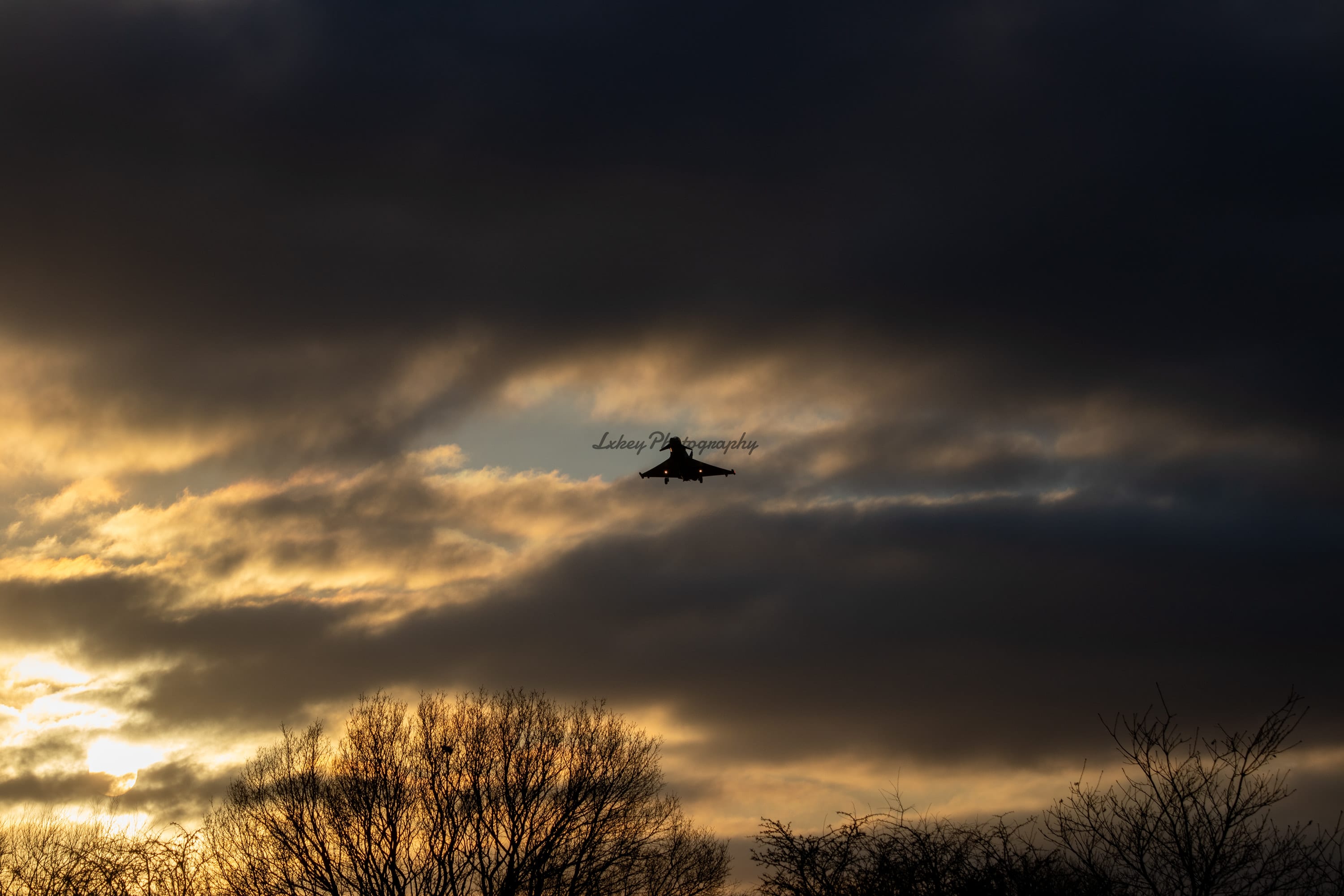 Fast jet silhouetted against a break of golden cloud at dusk above a treeline