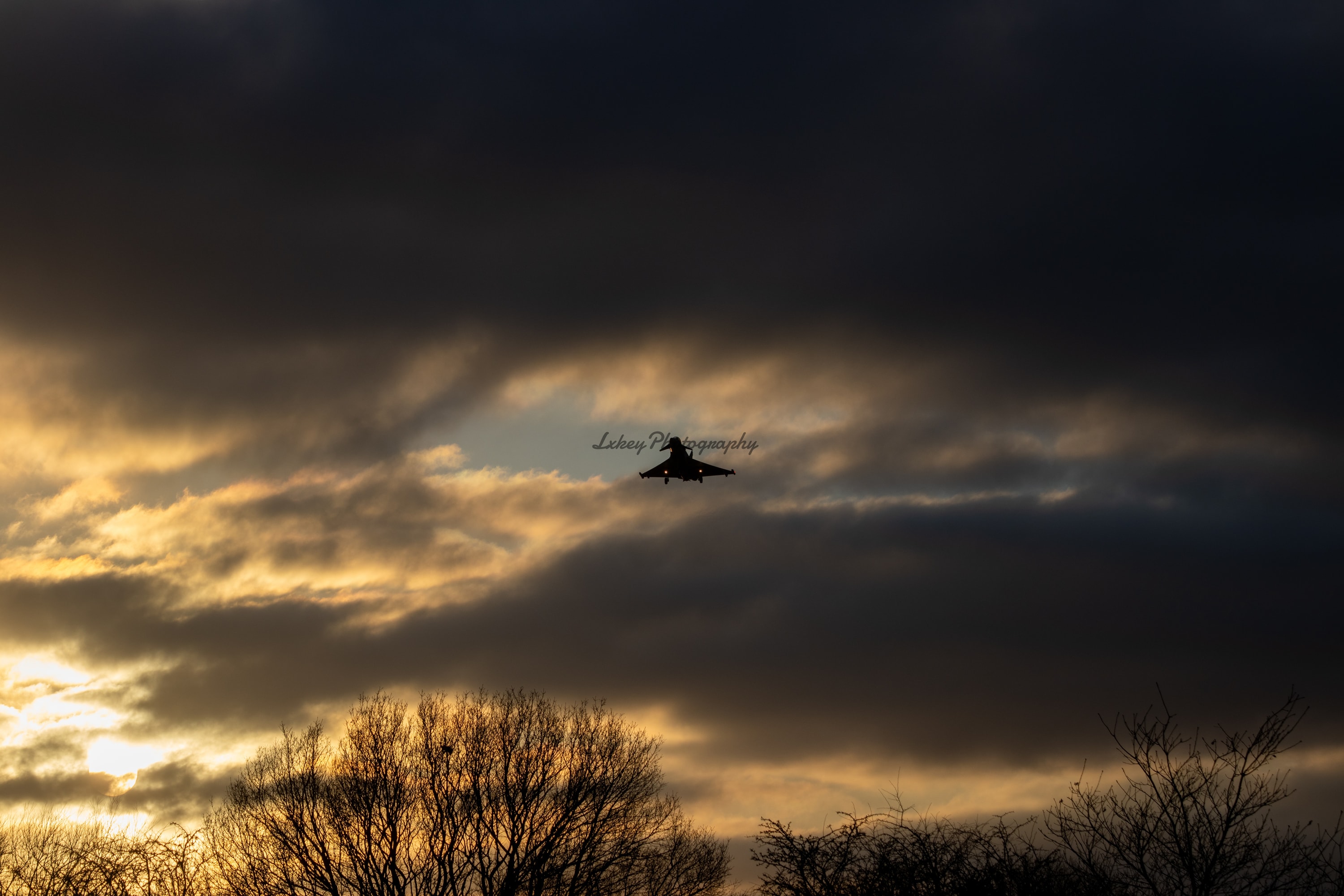 Fast jet silhouetted against a break of golden cloud at dusk above a treeline